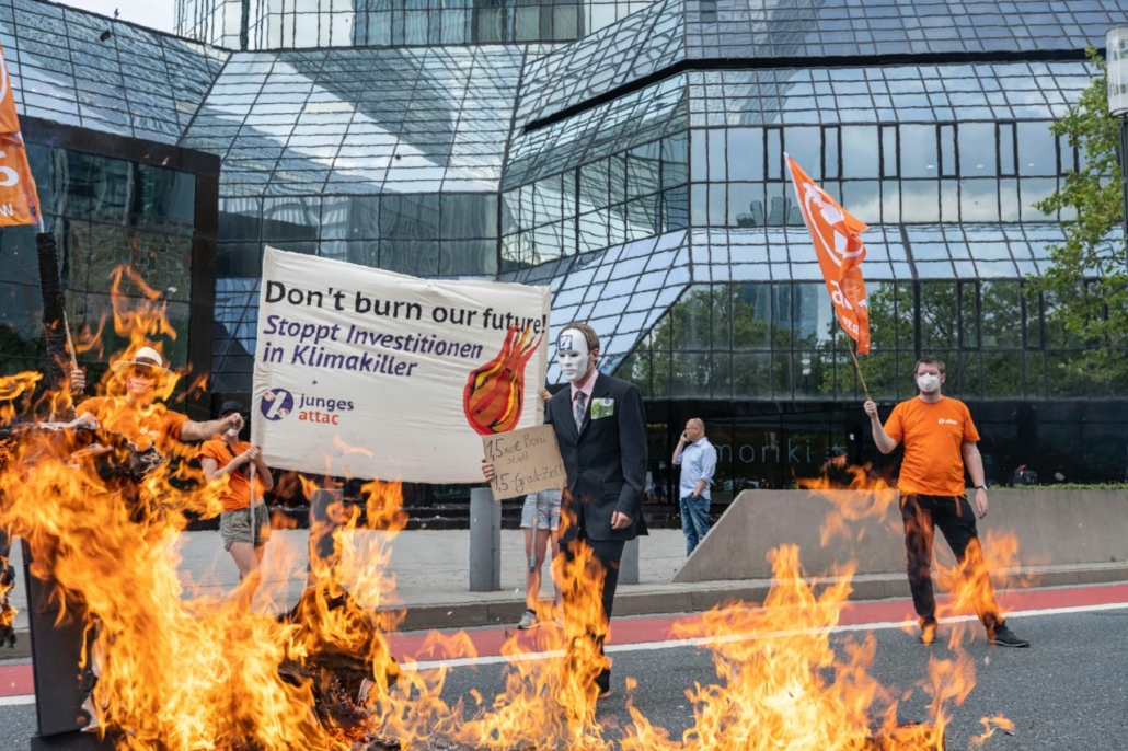 Im vordergrund sieht man Reste eines brennenden Deutsche-Bank-Logos, hinter den Flammen stehen mehrere Attacies mit einem Transparent auf dem "Don't burn our future – Stoppt Investitionen in Klimakiller" steht, im Hintergrund ist die Deutsche-Bank-Zentrale