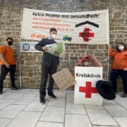Ein als Krankenhaus verkleideter Mensch bittet um Spenden um die Löhne bezahlen zu können, während ein als Krankenhausmanager verkleideter Mensch mit 100-Euro-Scheinen wedelt. Im Hintergrund ist das Transparent mit dem Slogan "Keine Profite mit Gesundheit" zu sehen