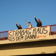 An einer Brücke hängt ein Transparent mit dem Slogan "Strabag raus aus dem Danni", dahinter recken drei Aktivist*innen ihre Faust in den Himmel.