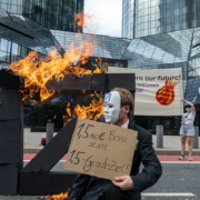 Ein als Banker verkleideter Aktivist steht vor dem brennenden Logo und hält ein Schild auf dem "1,5 Mio € Rendite statt 1,5 Grad" steht