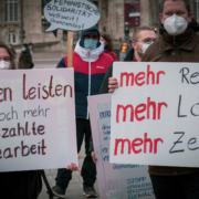 Auf der Kundgebung zum 8. März sieht man zwei Aktivist*innen die Schilder halten eins mit dem Slogan "Frauen leisten noch mehr unbezahlte Sorgearbeit" das andere hat den Slogan "mehr Respekt, mehr Lohn, mehr Zeit"