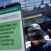 Vor dem Darmstadtium steht ein als Polizist mit Helm verkleideter Mensch, der ein Schild, das ein überdimensioniertes Handy mit einem fiktiven Whats-App-Chat der Polizei Südhessen, darin ist die Nachticht "Willkommen zur Chatgruppe für Hakenkreuze, Hitlerbilder und Mobbing. Viel Spaß
