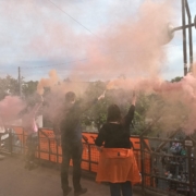 Auf einer Brücke stehen drei Menschen, die orangen Rauch hochhalten, vor ihnen hängt ein Transparent von der Brücke unter der die Demo entlaggeht