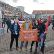 Drei junge Menschen stehen mit einer orangen Fahne auf der "Gemeinsam Nazis stoppen" steht auf einer Kundgebung