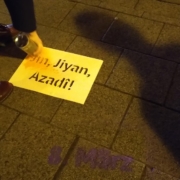 Eine Hand sprüht mit oranger Sprühkreide ein Stencil mit dem Slogan "Jin, Jiyan, Azadî! aus
