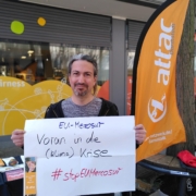 Ein Mensch hält an einem Infostand mit Attac-Beach-Flag ein Schild auf dem Mit Edding geschrieben ist "EU-Mercosur Voran in die (Klima) Krise #stopEUMercosur“