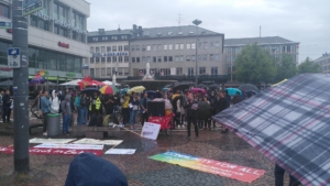 Menschen mit Regenschirmen stehen vesammelt um auf den Bodenliegende Transpis.