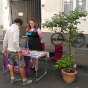 Eine Frau steht hinter einem Infostand von junges Attac auf dem viele Sticker liegen, davor schaut sich ein Mensch die Auslage an.