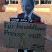 Vor einen Pappaufsteller von Christian Lindner hält jemand ein Schild mit dem Text "Für bezahlbare Preise von Energie, Wohnen, Mobilität"