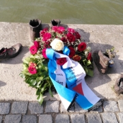 Ein Kranz aus Rosen mit einem weiß-blauen Band mit der Aufschrift: "Zum ehrenden Gedenken, Bund der Antifaschistinnen und Antifaschisten liegt zwischen einem Denkmal aus bronzenen Schuhen. Dahinter sieht man das Wasser der Donau.