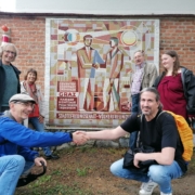 Zwei Attacies, einer aus Graz, einer aus Darmstadt geben sich vor einem Mosaik der Grazer Partnerstädte – auf dem sich zwei Männer die Hände geben – die Hände. Sie sind von Attacies aus Darmstadt und Graz umgeben.