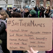 Ein Papschild mit der Aufschrift: "#SayTheirNames Ferhat Unvar, Said Nesar Hashemi, Hamza Kurtović, Vili Viorel Păun, Mercedes Kierpacz, Fatih Saraçoğlu, Sedat Gürbüz, Gökhan Gültekin" wird in die Kamera gehalten. Dahinter sind einige Menschen mit Papschildern zu sehen.