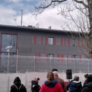 eine kleine Menschenmenge von hinten schaut über die Straße zu einem grauen Gebäude, dem Abschiebeknast.