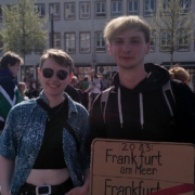 Zwei junge Menschen mit dem Schild 2083 Frankfurt am Meer und darunter durchgestrichen Frankfurt am Main