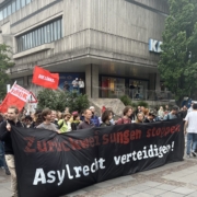 Im Vordergund ist ein großes, schwarzes Transparent zu sehen mit der Aufschrift: Zurückweisungen stoppen - Asylrecht verteidigen! Dahinter ist der Beginn des Demozugs zusehen mit verschiedenen Fahnen. Gut zu sehen sind die Fahnen der Linken und der Solid.