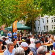 Viele Menschen an Tischen oder stehend sich unterhaltend. Im Hintergrund sieht man den Attac Pavillion