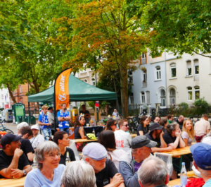 links-buntes Sommerfest Viele Menschen an Tischen oder stehend sich unterhaltend. Im Hintergrund sieht man den Attac Pavillion