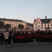 Menschenmenge mit Fahnen auf dem Marktplatz. Im Fordergrund ist ein Schwarzes Transpi mit der Aufschrift "Siamo Tutti Antifascisti" zu sehen