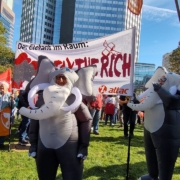 Im Fordergrund steht ein als Elefant verkleideter Mensch, dahinter hält eine weitere Person im Elefantenkostüm und eine Mit Attac Fahne ein Transpi mit der Aufschrift: Der elefant im Raum: Tax the rich